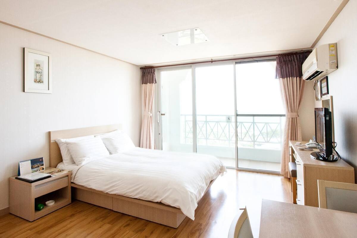 A hotel room with a bed, a bedside table, a desk with a chair, a TV, and a balcony with curtains.