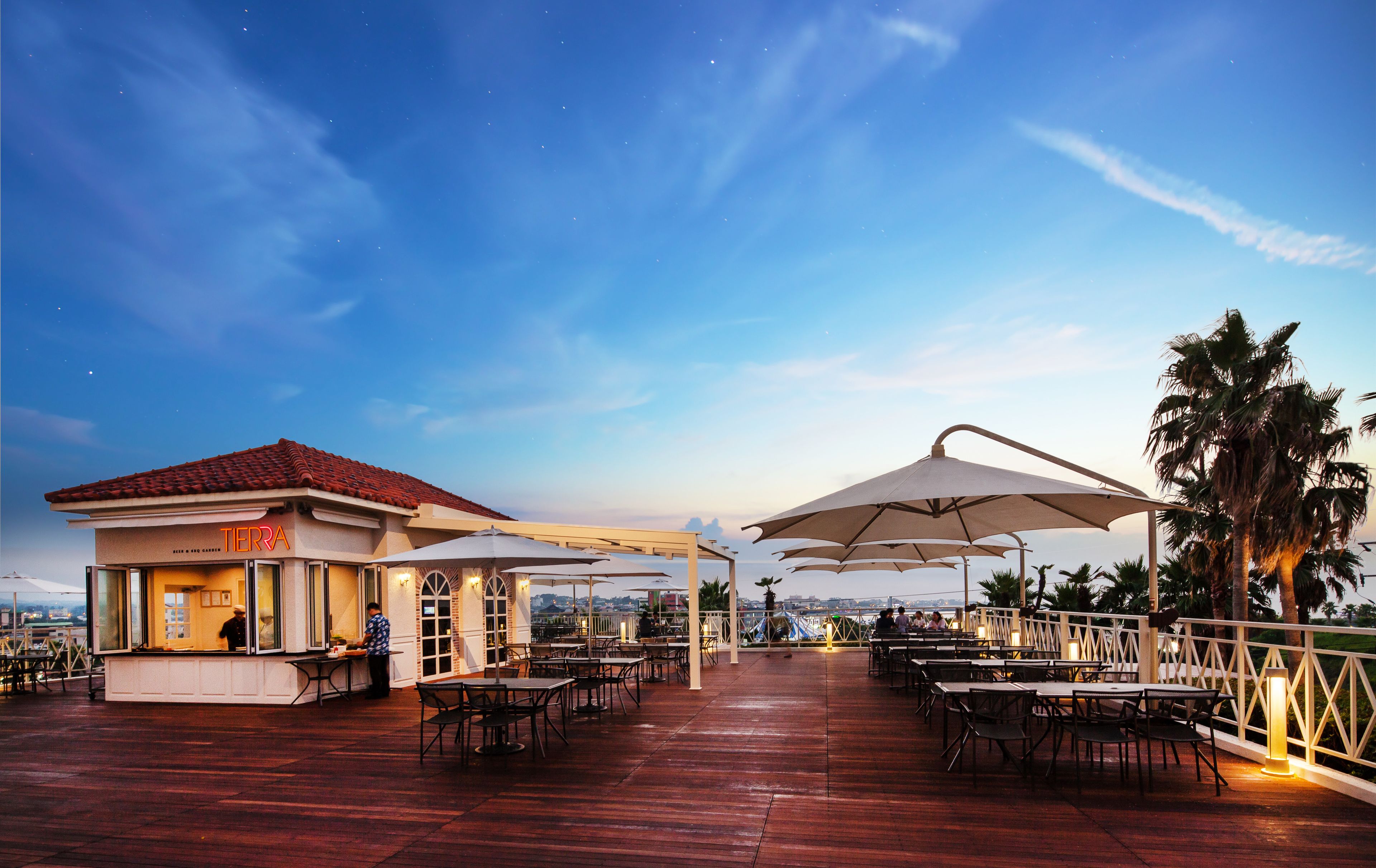 An outdoor restaurant with tables and umbrellas, a bar area, and a view of the city skyline.