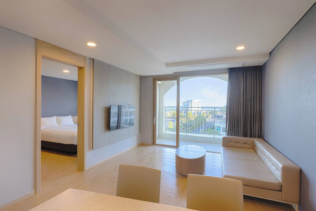 A modern hotel room with a bed, a sofa, a dining table, and a balcony with a view of the city.
