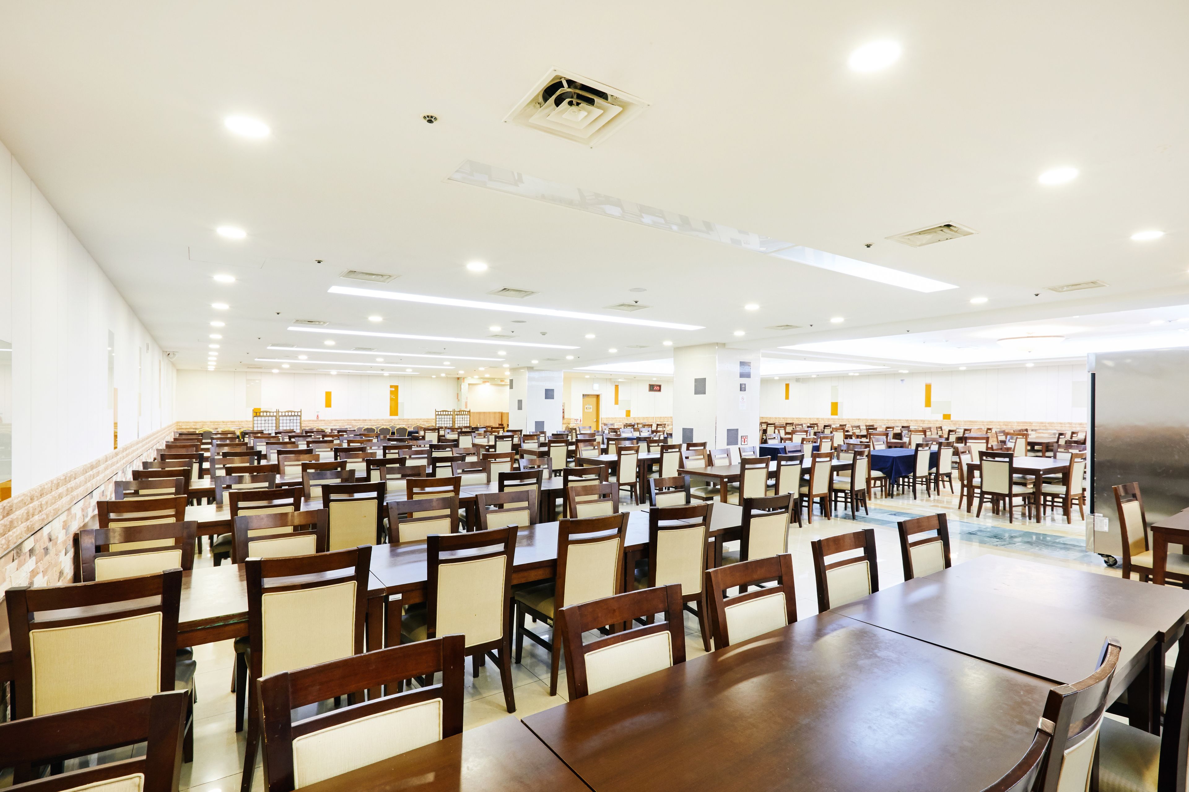 A large dining hall with rows of tables and chairs, high ceilings with recessed lighting, and a clean, modern design.