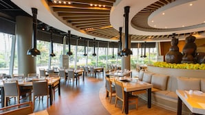 Un restaurant spacieux avec de hauts plafonds, un éclairage moderne suspendu et un espace de repos central agrémenté d’une composition florale décorative.
