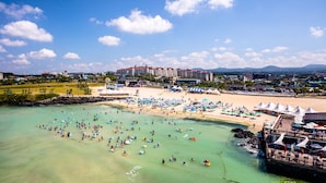 Vue aérienne d’un complexe hôtelier en bord de mer, avec plusieurs bâtiments, une plage de sable et de nombreuses personnes pratiquant des activités nautiques.