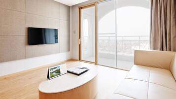 Ein modernes Wohnzimmer mit einem Flachbildfernseher, einem runden Couchtisch mit Kalender und Notizbuch, einem beige farbenen Sofa und einer Glastür, die auf einen Balkon führt.