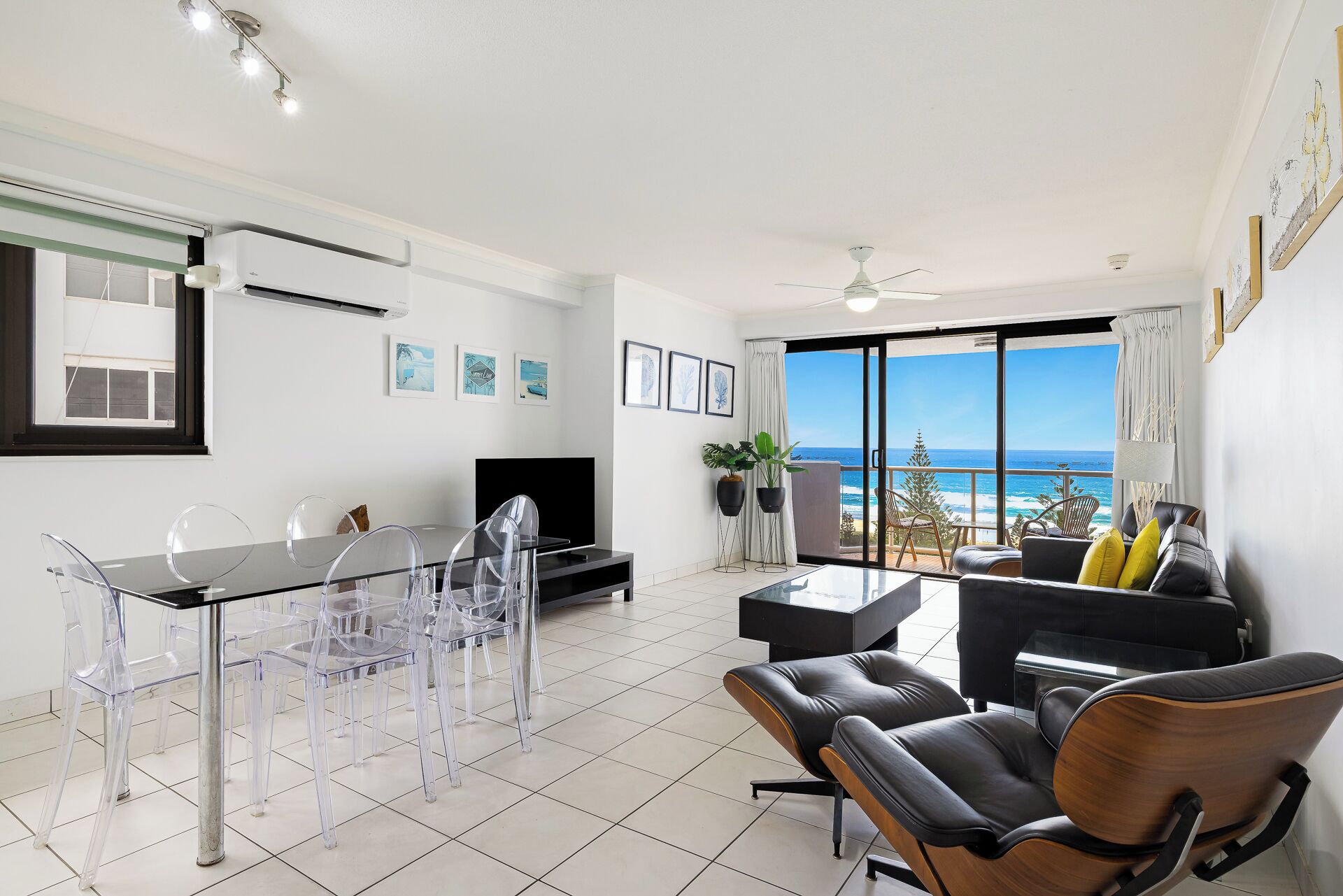 Una sala de estar moderna con un amplio ventanal con vista al océano, una zona de comedor con sillas traslúcidas y un televisor de pantalla plana.