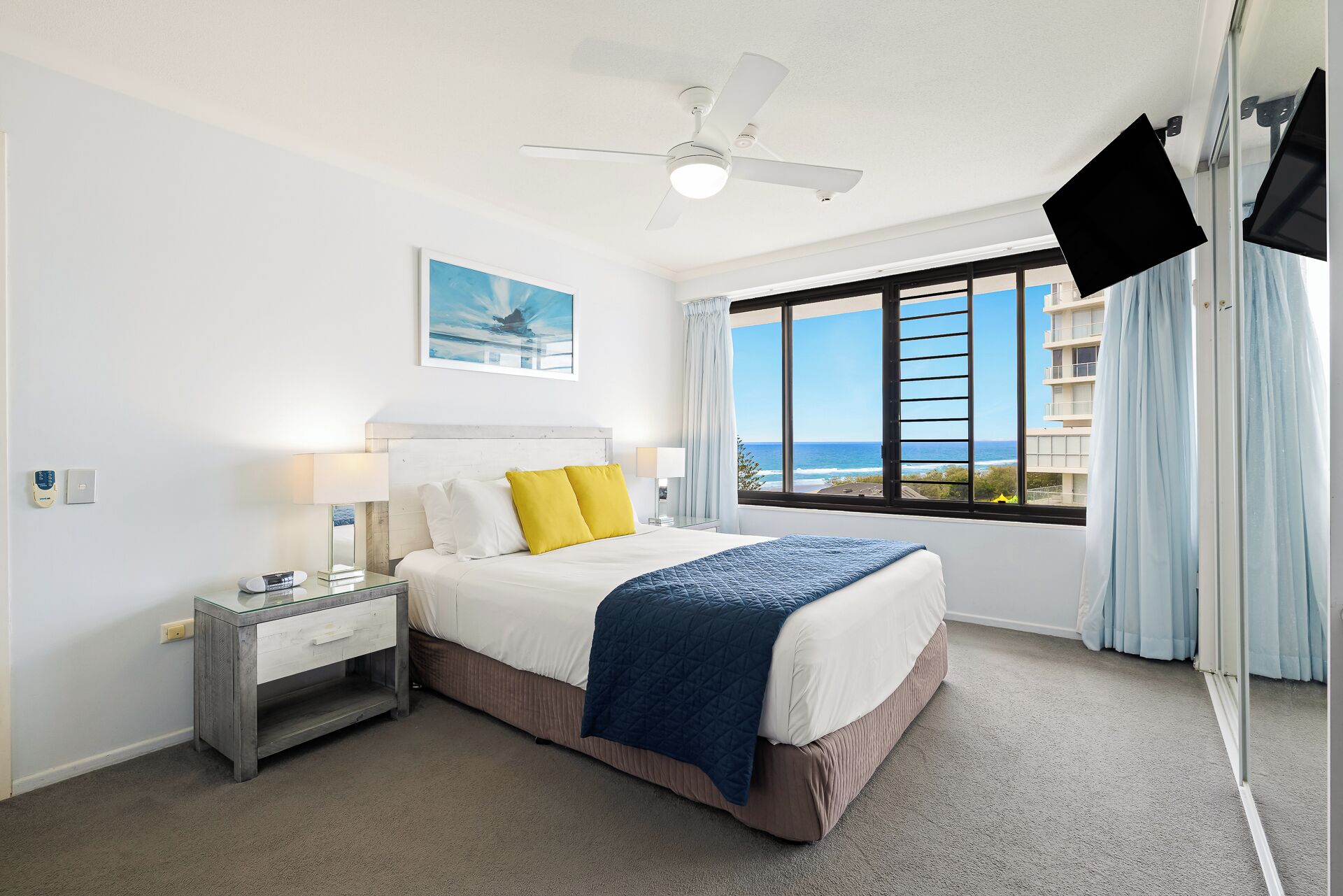 A hotel room with a bed, a nightstand, a desk, a ceiling fan, a TV, a window with a view of the ocean, and a painting on the wall.