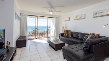 A modern living room with a black leather sofa, a glass coffee table, and a television on a wooden stand. There is a balcony with a view of the sea.