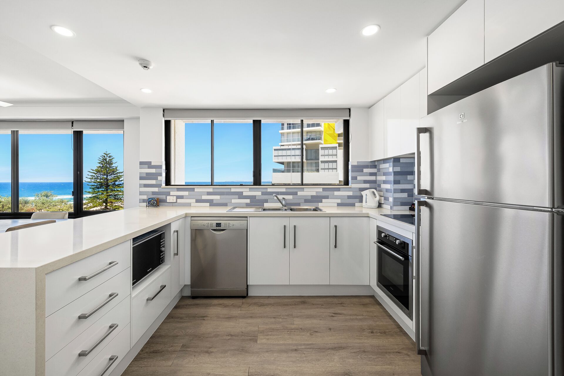 A modern kitchen with a dishwasher, oven, and stainless steel refrigerator. It features a large window with a view of the ocean and a tiled backsplash.