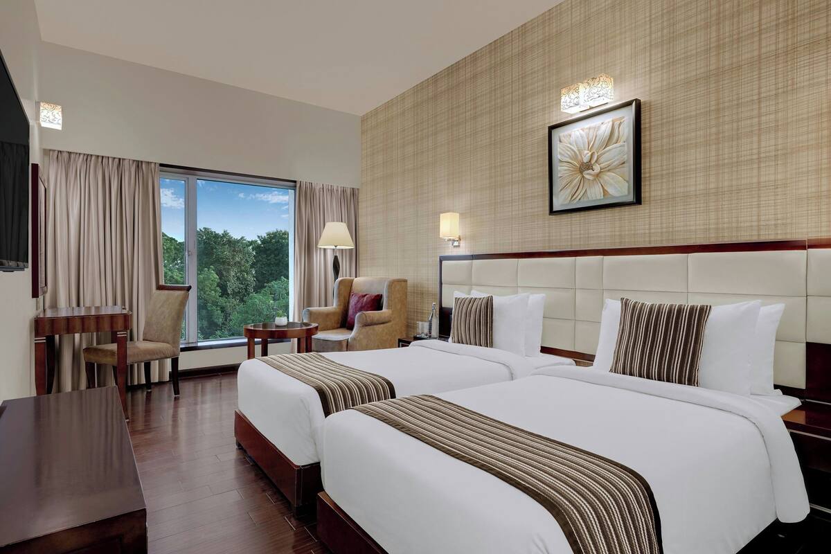 A hotel room with two beds, a desk, a chair, a lamp, and a window with curtains.