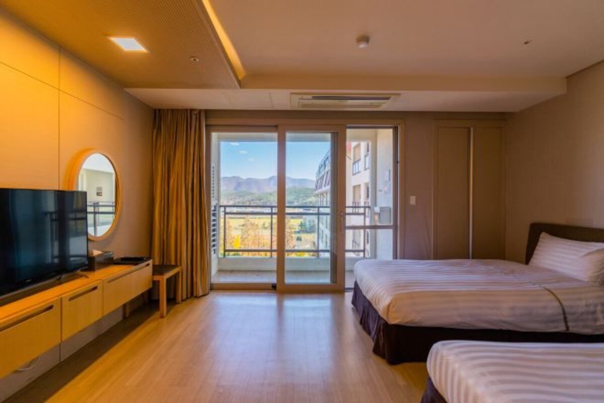A hotel room with a large bed, a TV, a small table, and a view of the city through a sliding door.