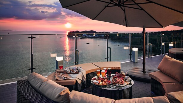 A sunset view from a balcony with outdoor seating, a table with food and drinks, and a pool in the background. - Sono Belle Byeonsan (Buan)