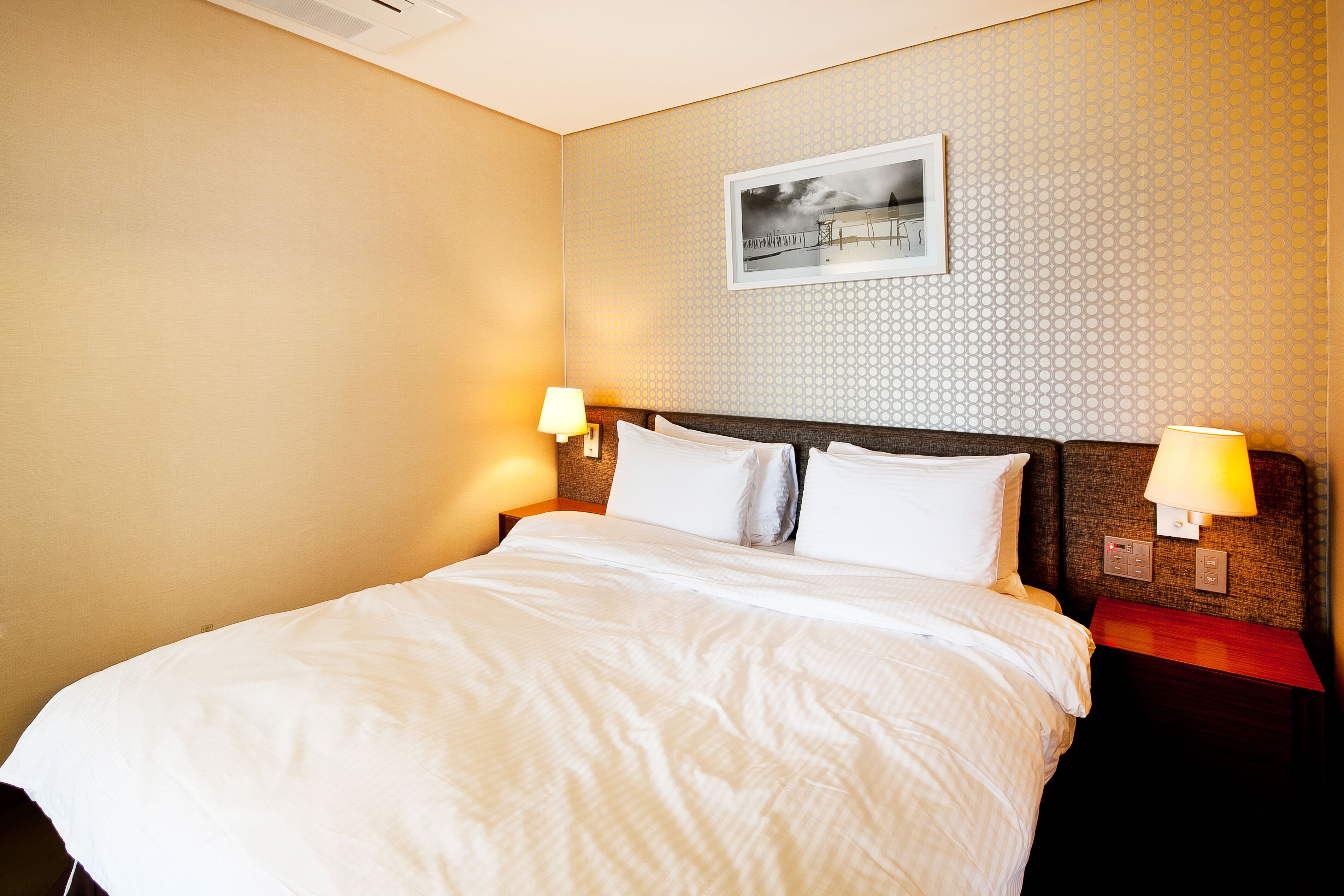 Habitación de hotel con una cama grande, dos mesitas de noche con lámparas, un cuadro en la pared y una pared con textura.