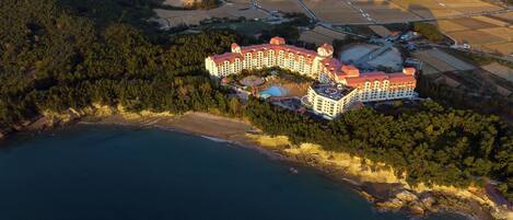 An aerial view of a coastal hotel with a beachfront, surrounded by dense forest and agricultural fields.