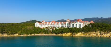 A large hotel with multiple floors and a red roof, situated by a body of water with a forested hillside in the background.
