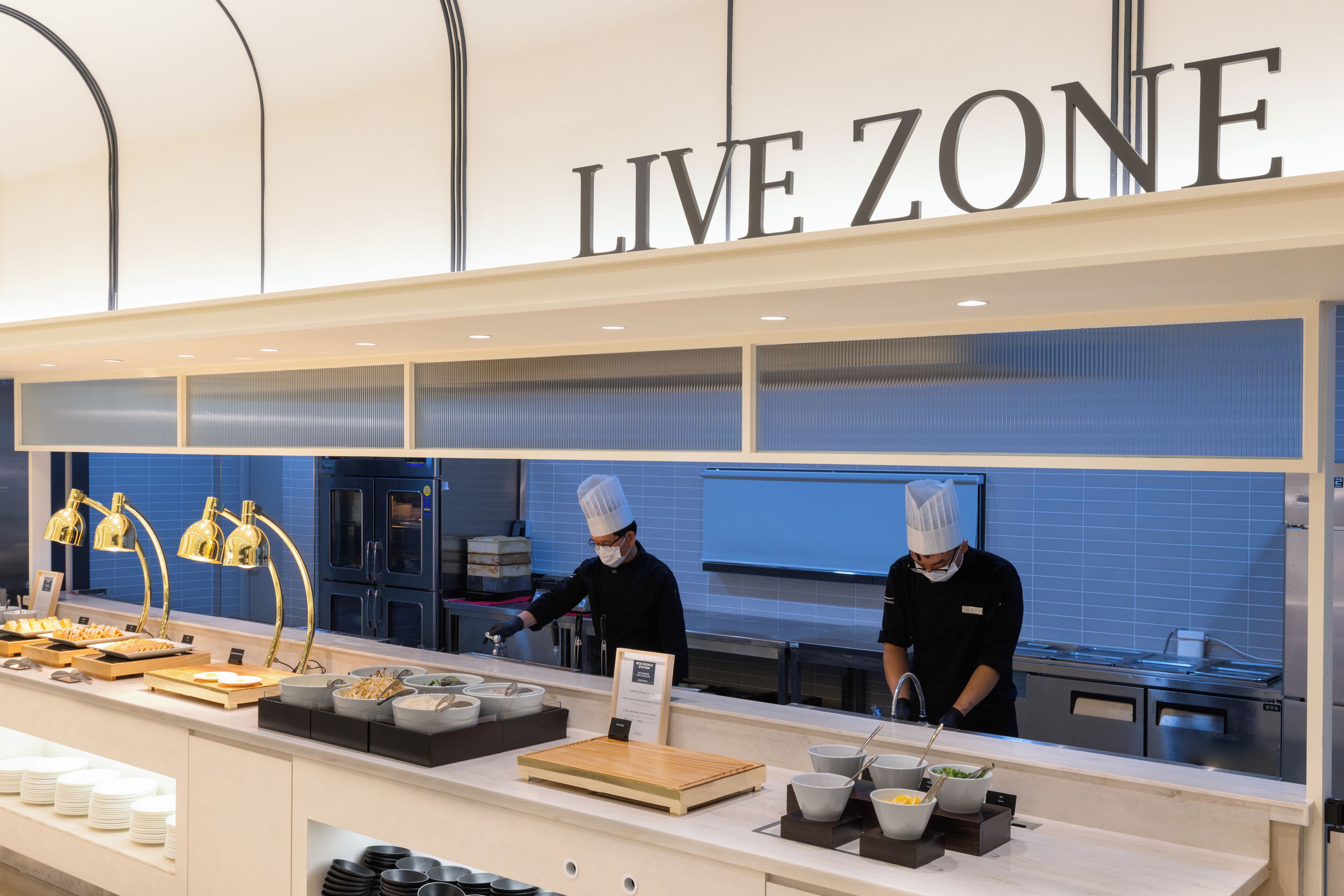 A kitchen area with the sign 'LIVE ZONE' above, two chefs in uniform, and various food items and utensils on the counter.