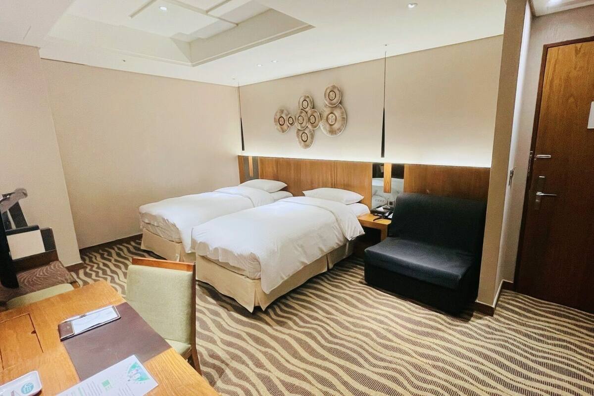 Habitación de hotel con dos camas, un escritorio, una silla, un armario de madera y una lámpara de pared.