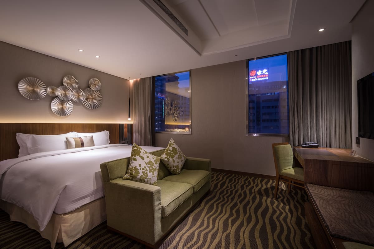 Habitación de hotel con cama, sofá verde, escritorio y una ventana grande con vistas a un paisaje urbano.