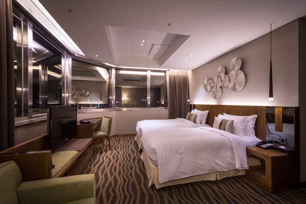 Habitación de hotel con una cama grande, un escritorio con computadora, una silla y vista de la ciudad a través de la ventana.