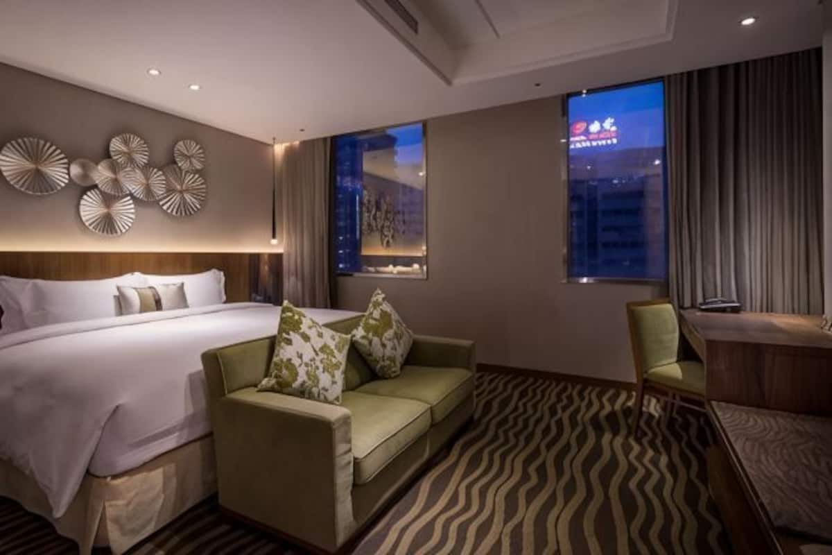 A hotel room with a bed, a green sofa, a desk, and a view of the cityscape.