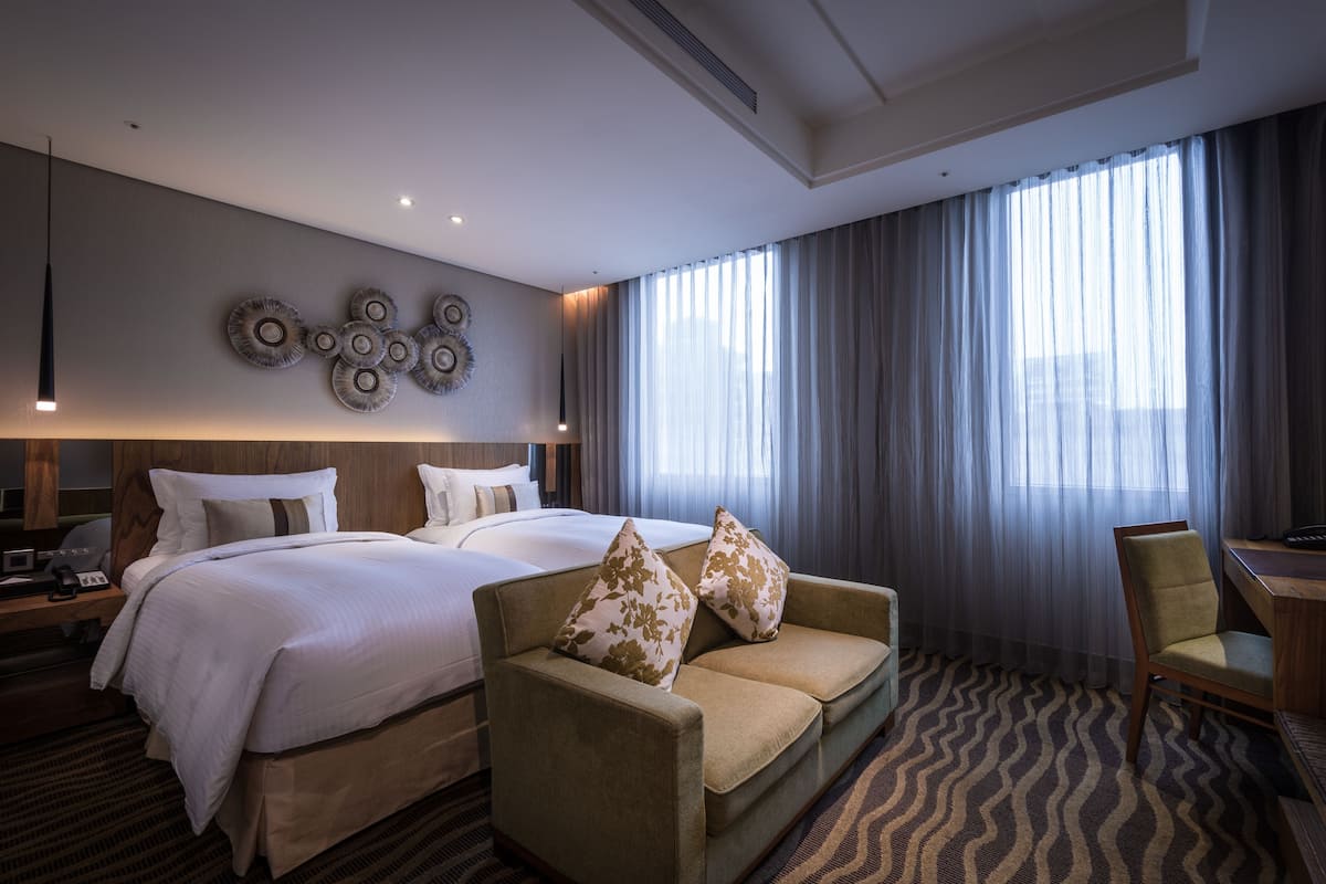 Habitación de hotel con una cama grande, un sofá, un escritorio y una silla. Se aprecia un ventanal con cortinas y un adorno en la pared sobre la cama.