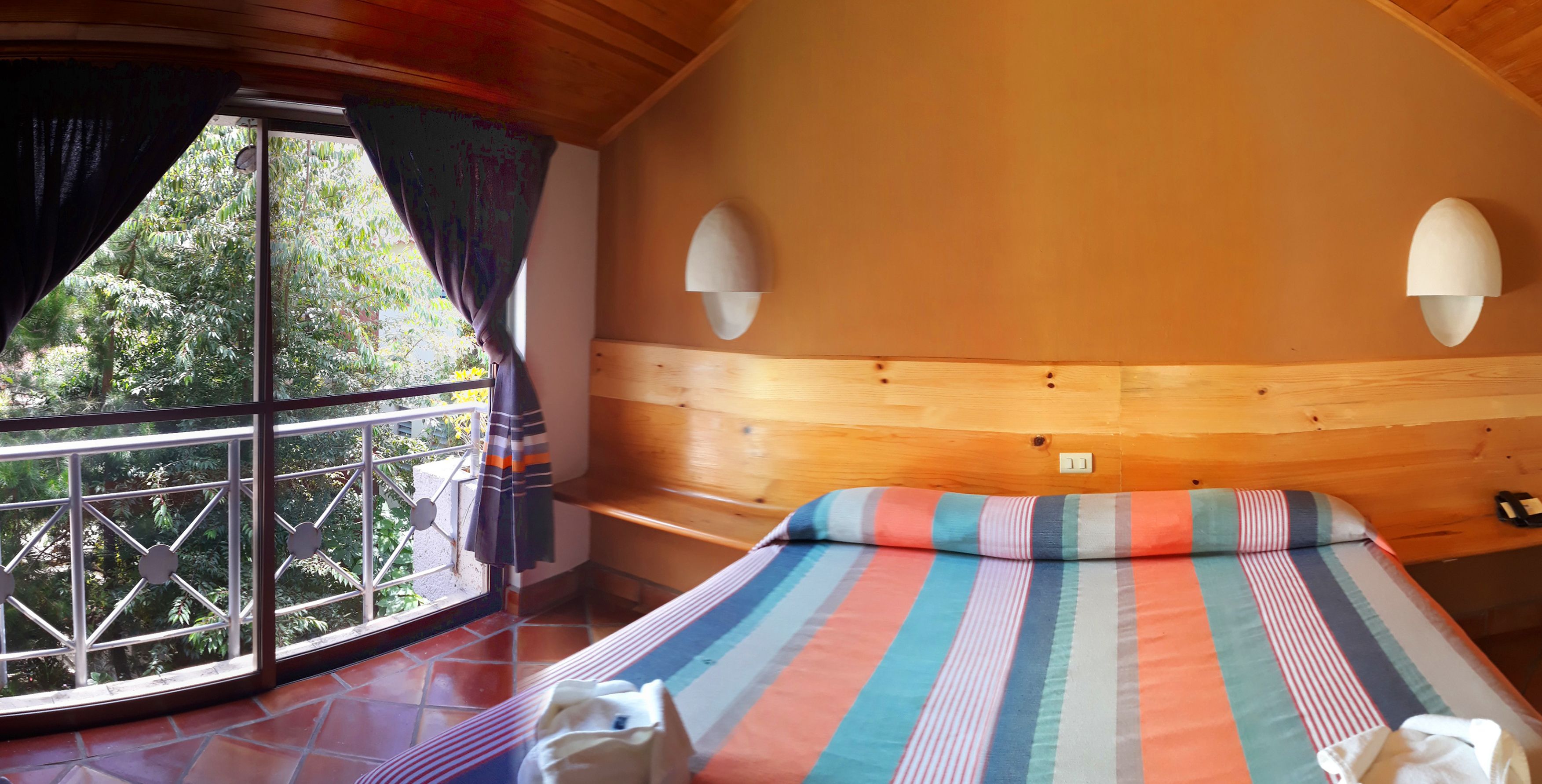 A room with a large window, a bed with striped bedding, wooden paneling, and two wall-mounted lamps.