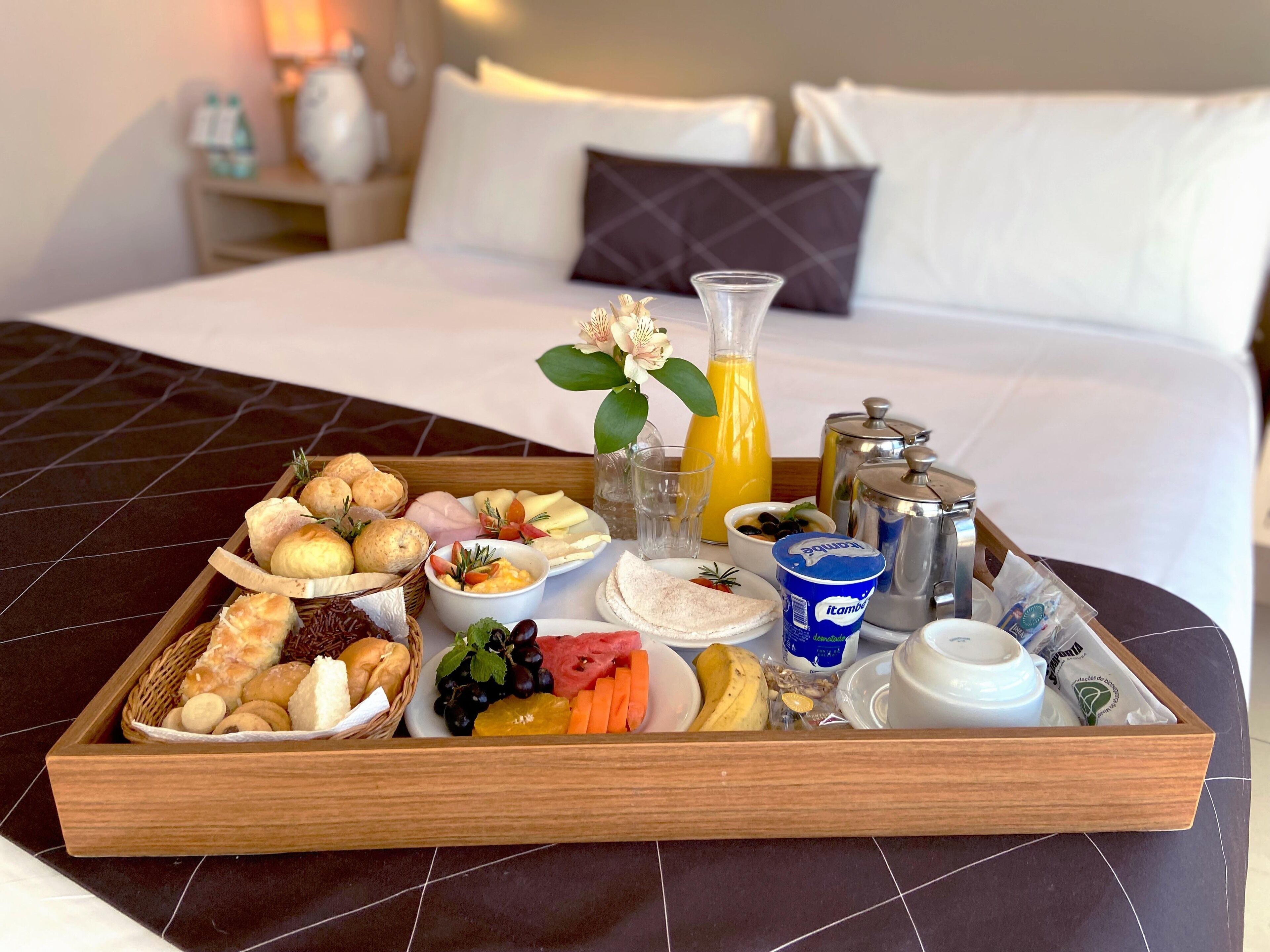 Una bandeja de desayuno sobre la cama, con pan, bollería, frutas, una botella de zumo de naranja, un vaso de agua, una cafetera y un recipiente de yogur.
