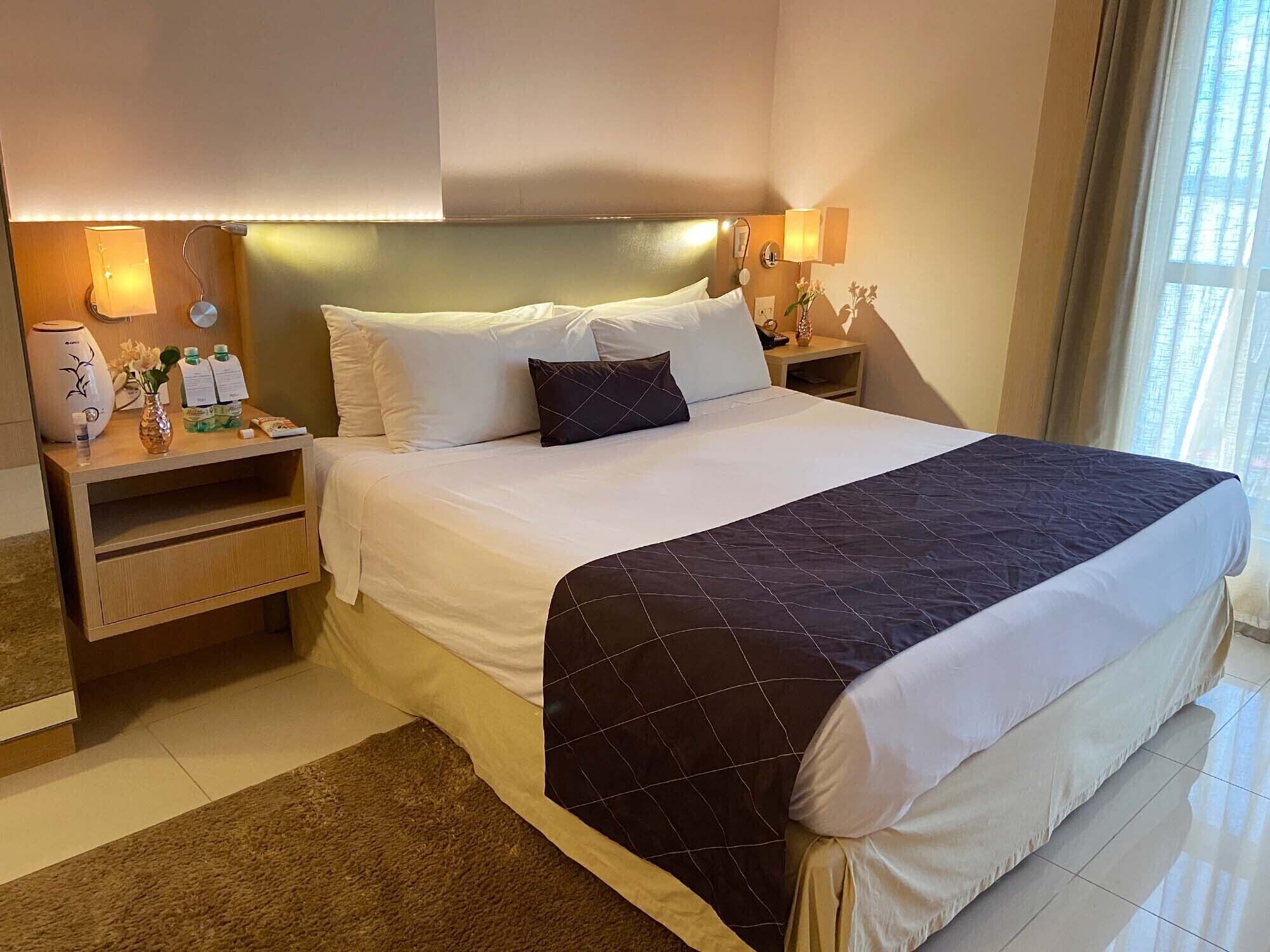 A hotel room with a large bed, two bedside tables, a nightstand, a vase with flowers, a lamp, and a window with curtains.