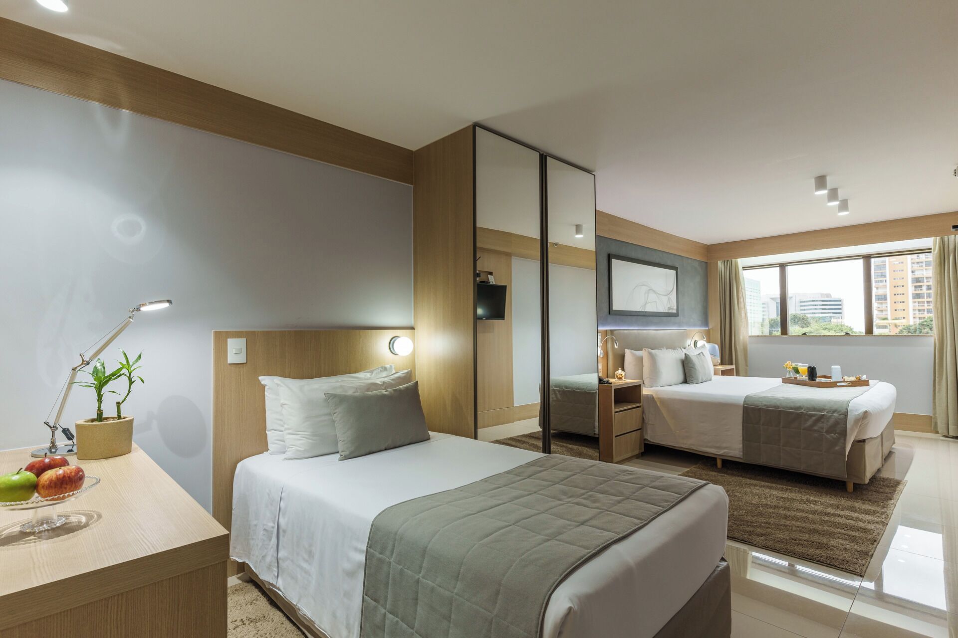 Quarto de hotel com duas camas, uma escrivaninha com abajur, um espelho e uma janela com vista para edifícios.