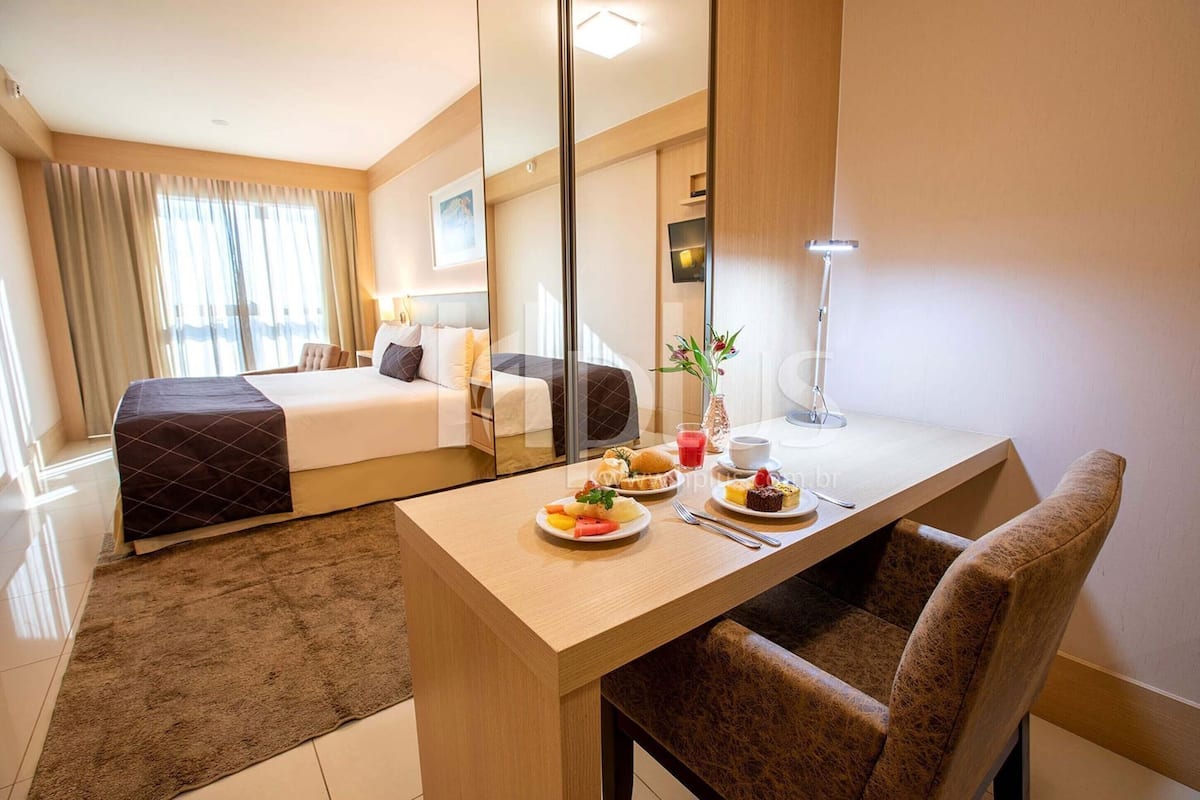 A hotel room with a bed, a desk with breakfast, a chair, and a large mirror.