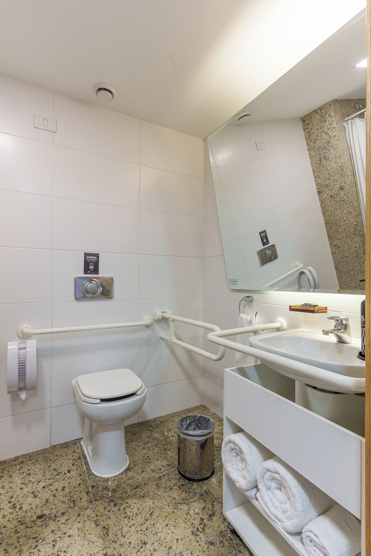 A bathroom with a toilet, grab bars, a sink, a trash can, and a towel rack.