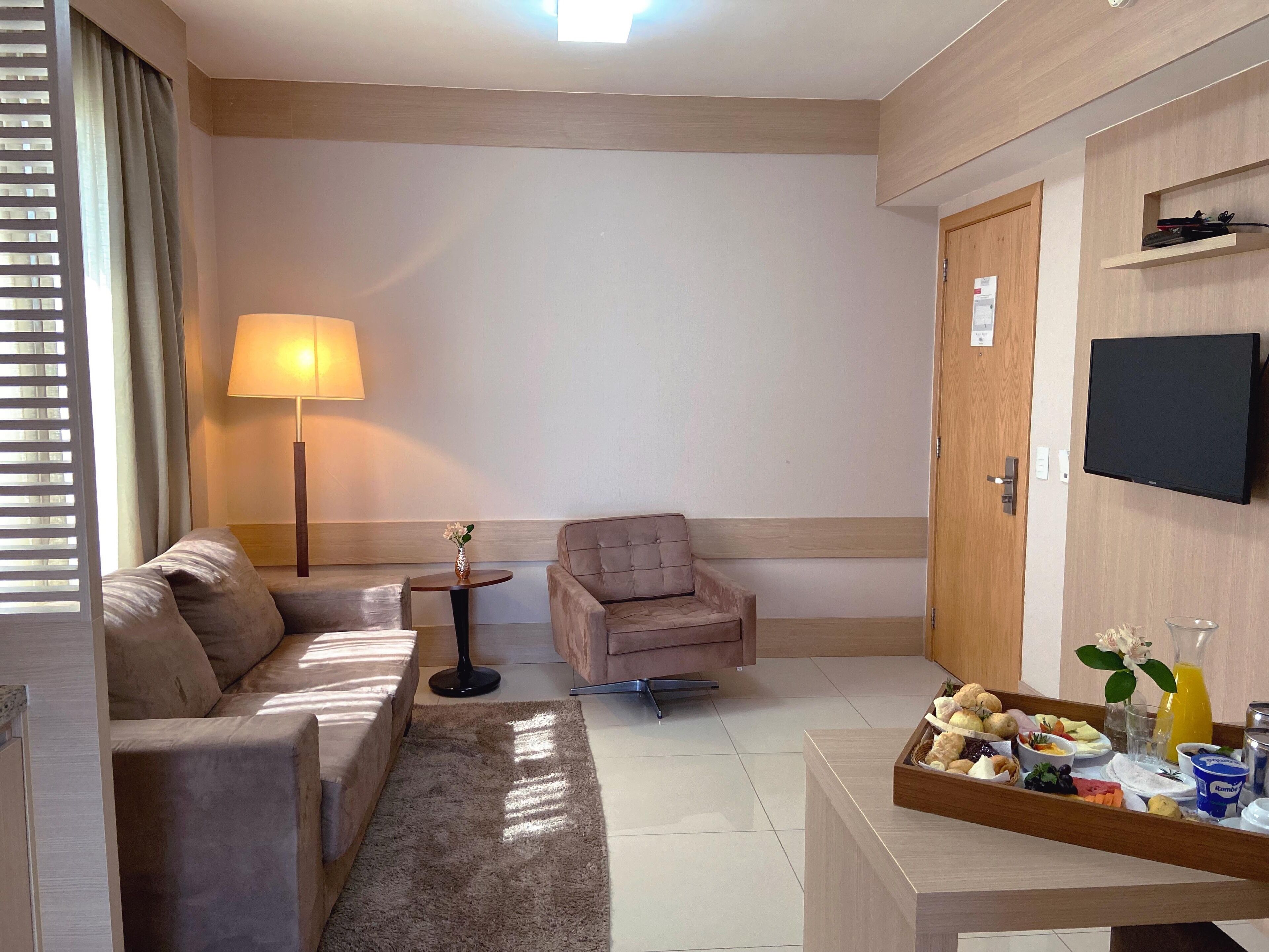 Una habitación de hotel moderna con sofá, sillón, una pequeña mesa con lámpara, televisión de pantalla plana y bandeja para el desayuno.