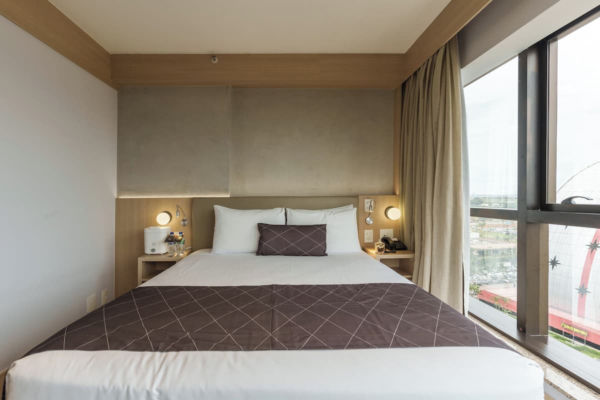 Una habitación de hotel con una cama grande, dos mesitas de noche con lámparas, vistas a la ciudad a través de amplios ventanales y un techo de madera.