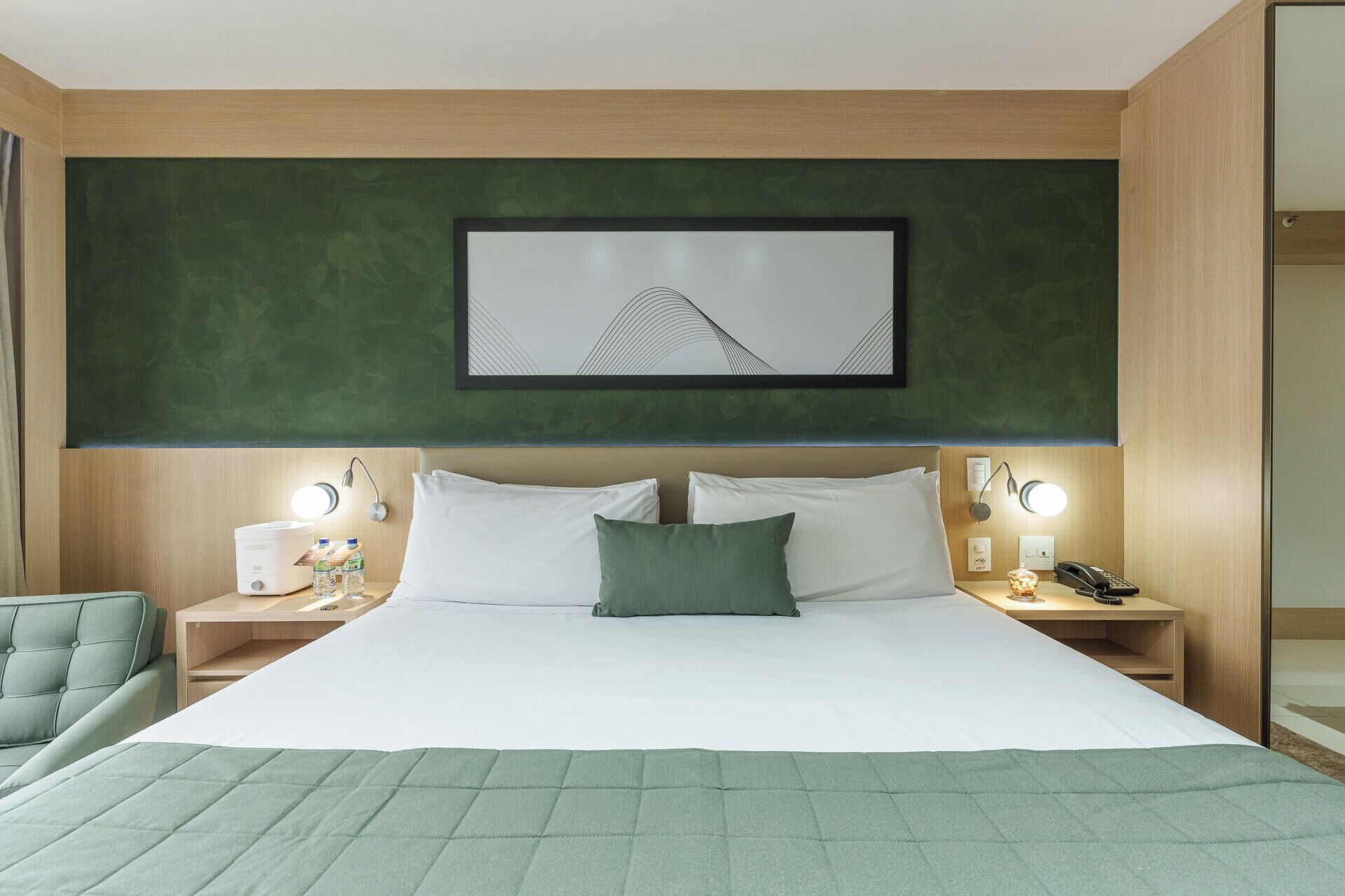 A neatly arranged hotel room with a large bed, two bedside tables, a green chair, and a framed artwork above the bed.