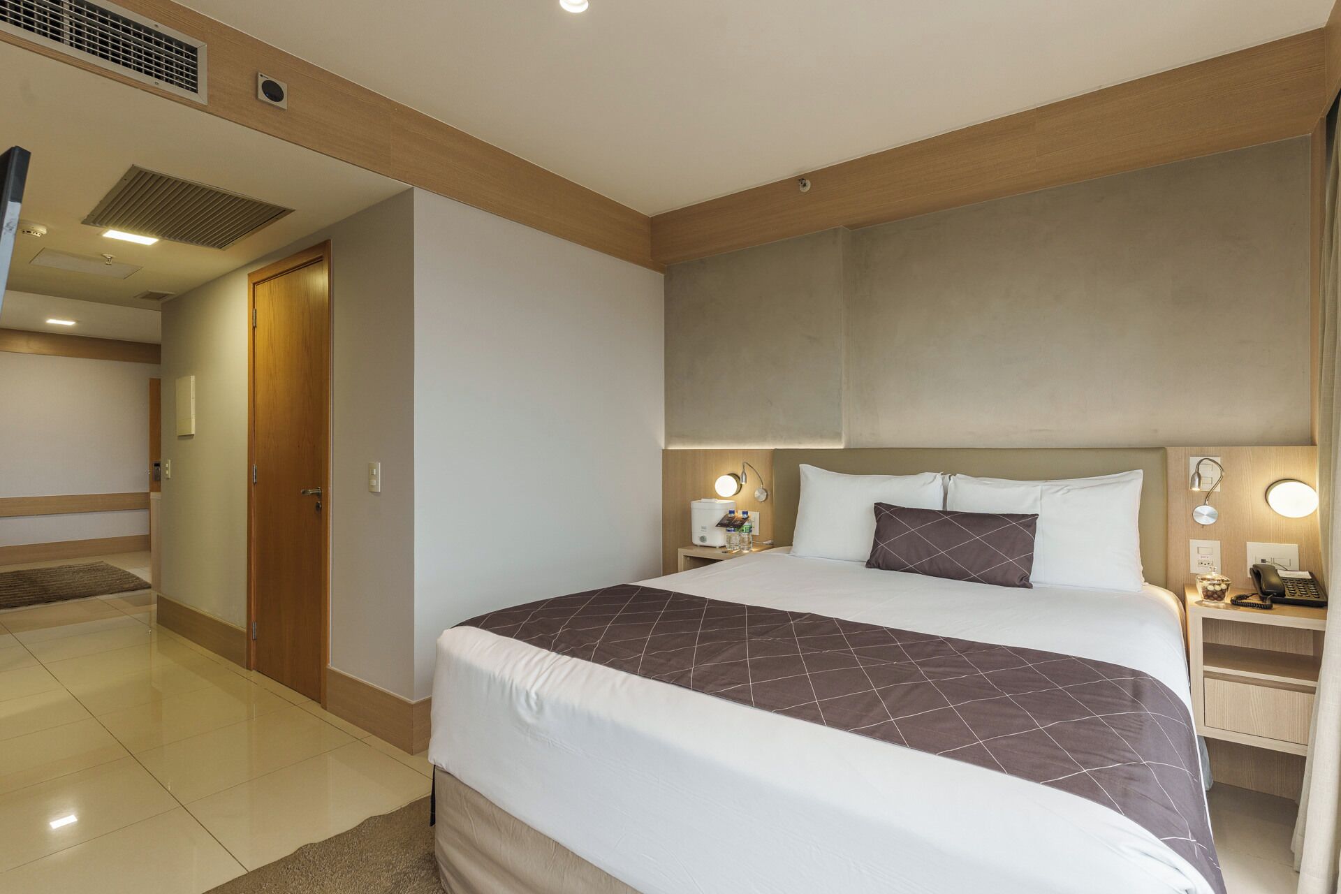 A hotel room with a large bed, bedside tables with lamps, a wooden headboard, and a door leading to another room.