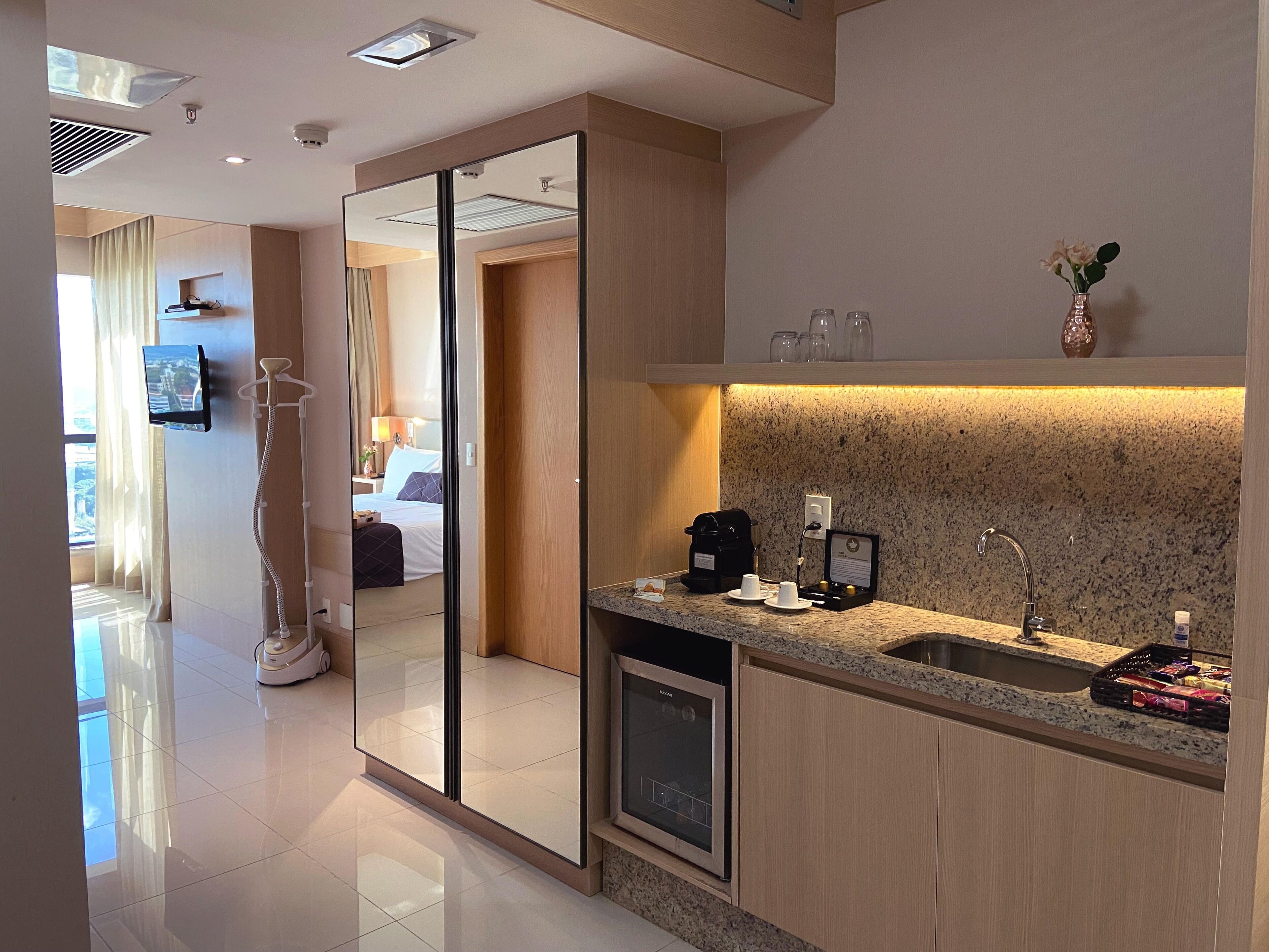 A modern hotel room with a large mirror, a kitchen area with a sink and coffee maker, and a view of the bedroom through the mirror.