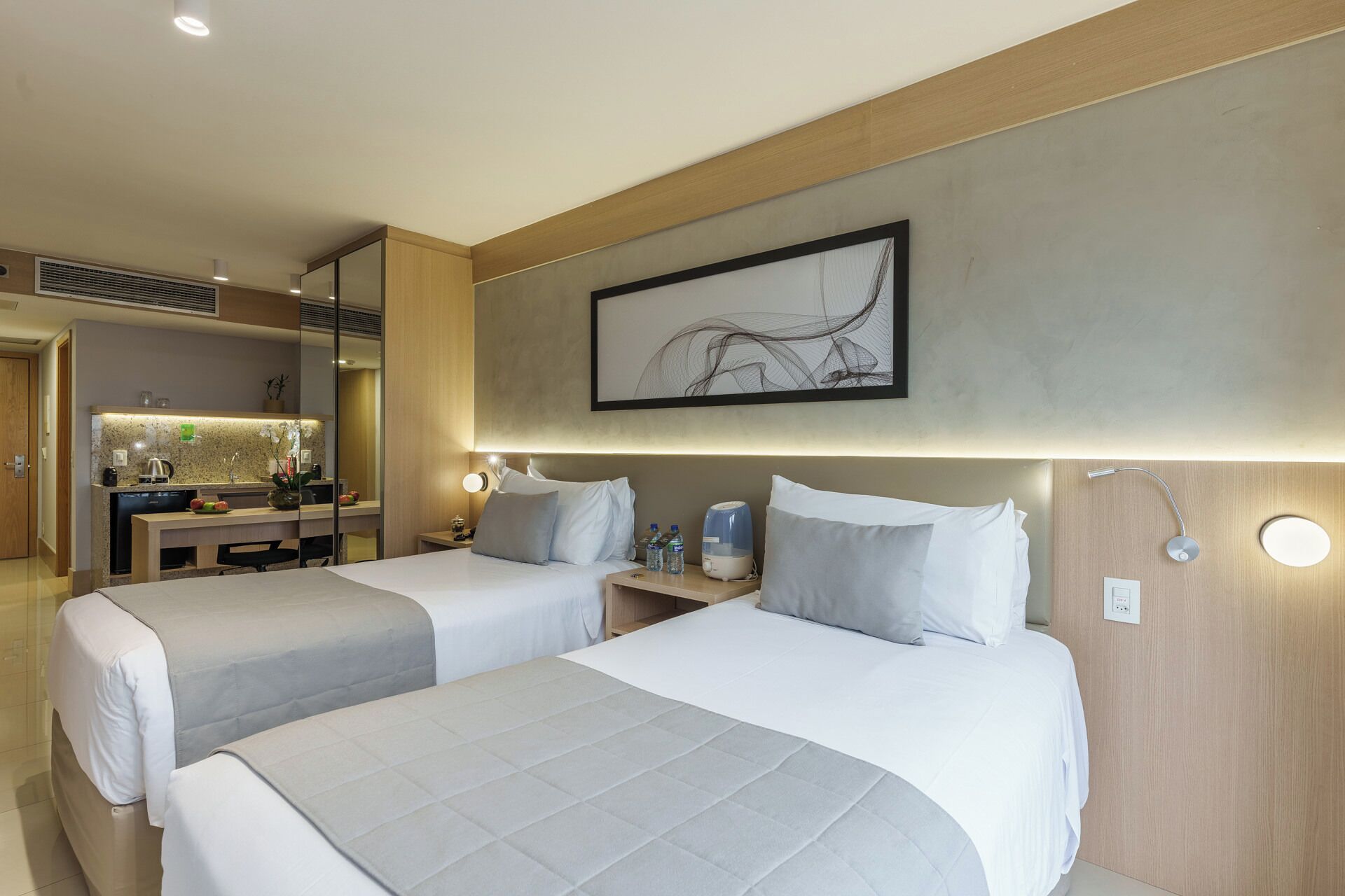 Quarto de hotel com duas camas, uma escrivaninha com cadeira, uma mesinha e uma obra de arte na parede.