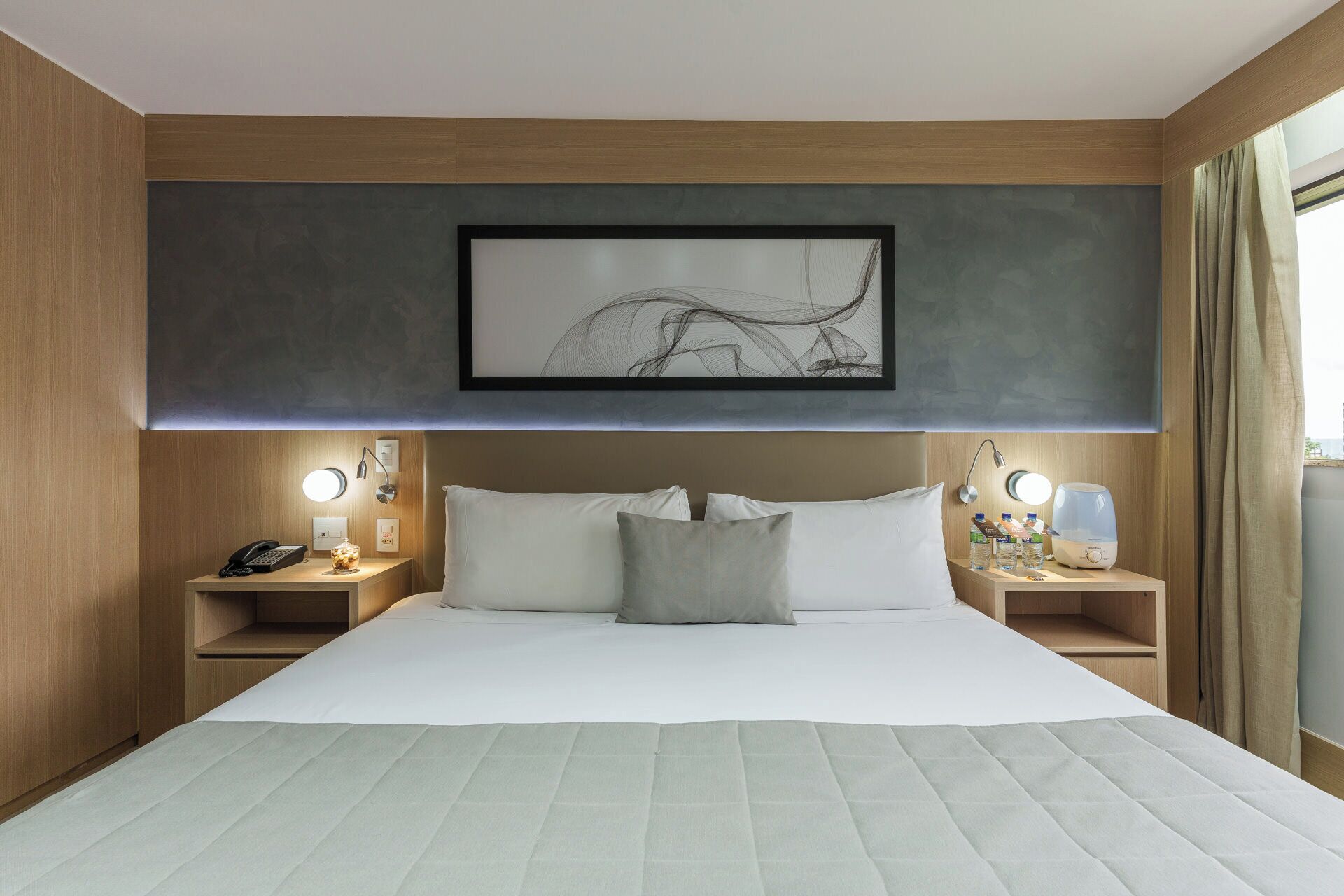A hotel room with a large bed, two bedside tables, a wall-mounted artwork, and a window with curtains.