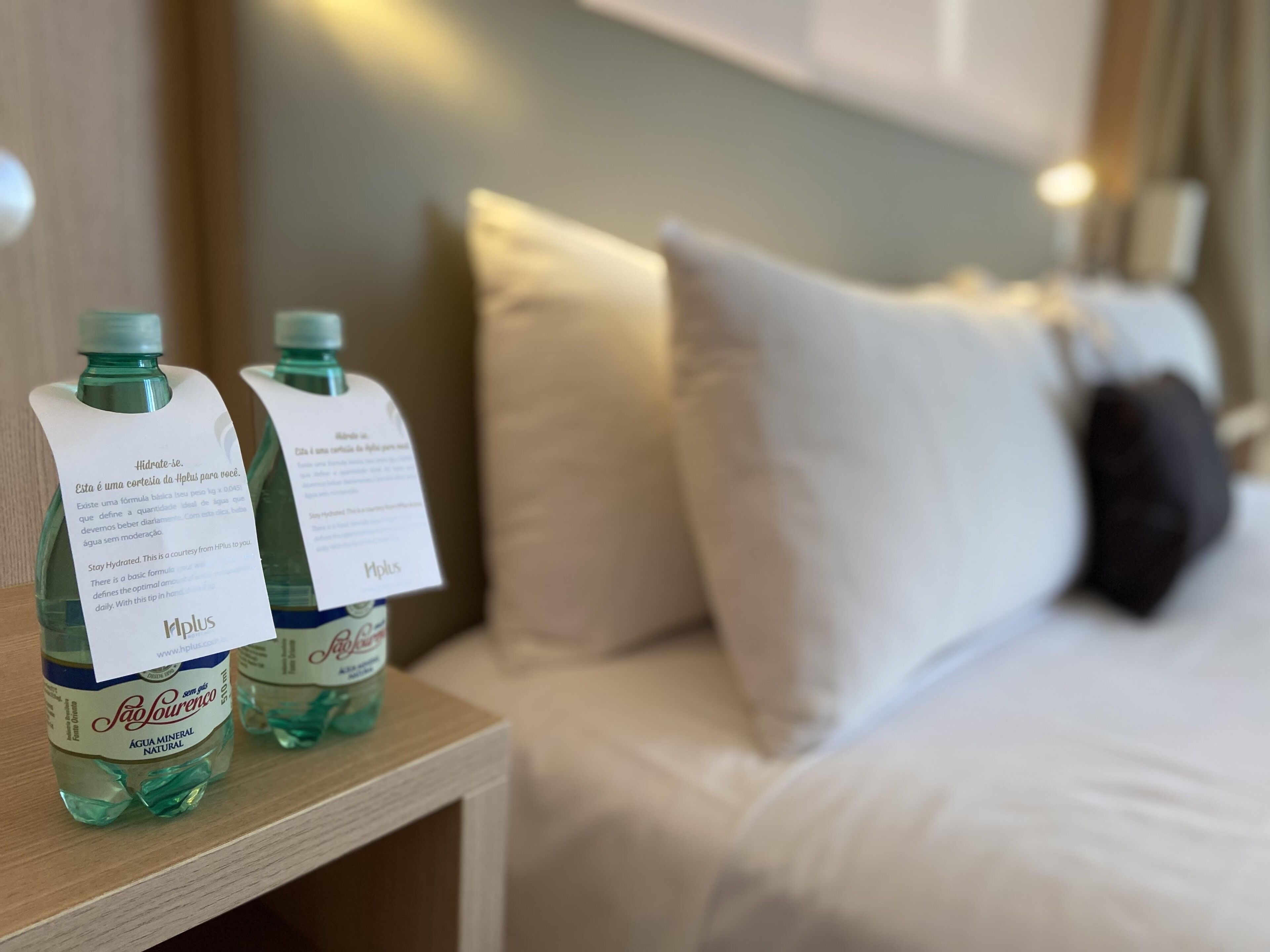 A hotel room with a bed, pillows, and two bottles of water with hydration reminders on the nightstand.
