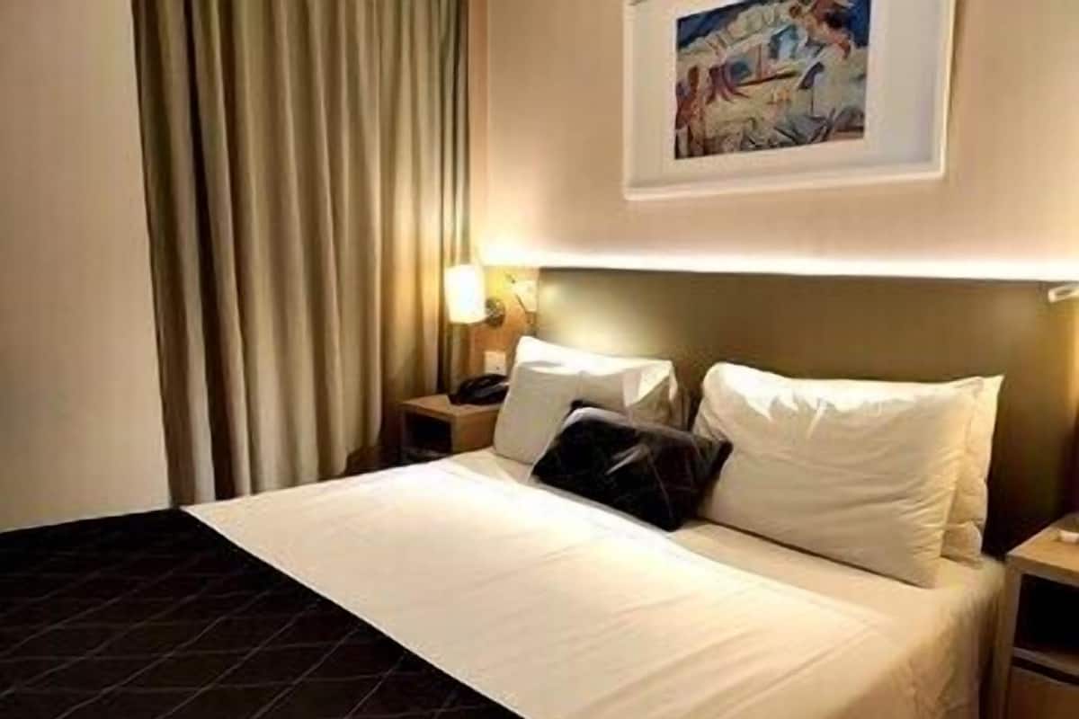 A hotel room with a bed, a nightstand, a lamp, a painting on the wall, and a window with curtains.