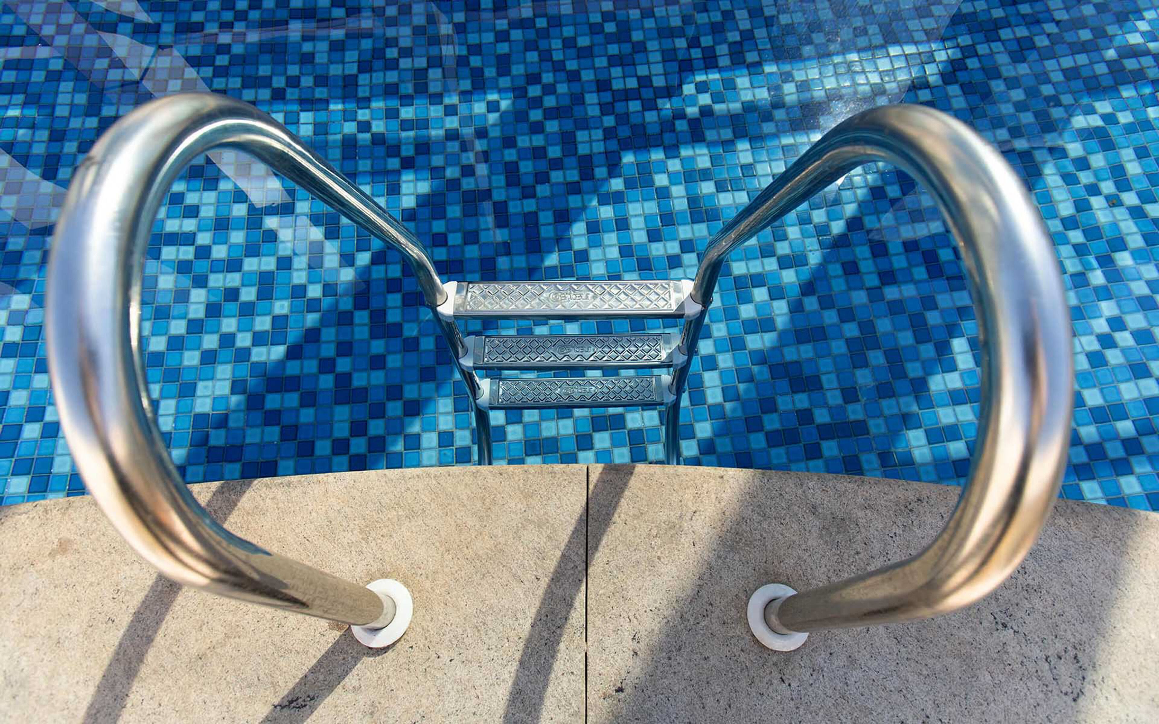 Escada de metal para piscina, com superfície antiderrapante, fixada em superfície de concreto, dando acesso a uma piscina com azulejos azuis em mosaico.