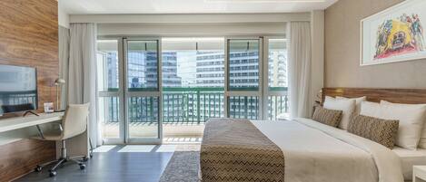 A modern hotel room with a large bed, a desk, and a view of the city through floor-to-ceiling windows.