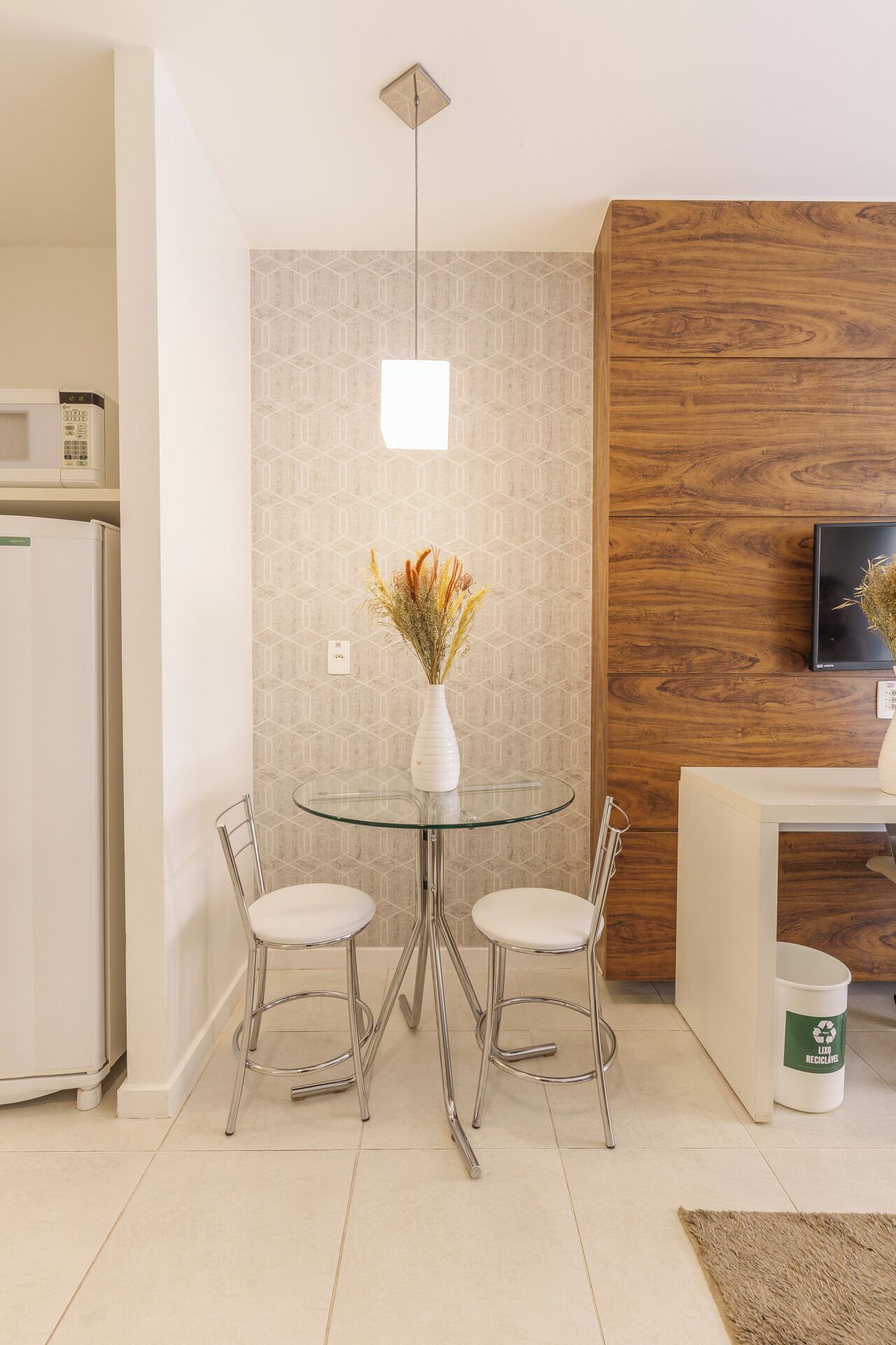 Una cocina moderna con una mesa redonda de vidrio, dos sillas, un florero blanco con flores secas, un microondas y una pared de acento de madera.