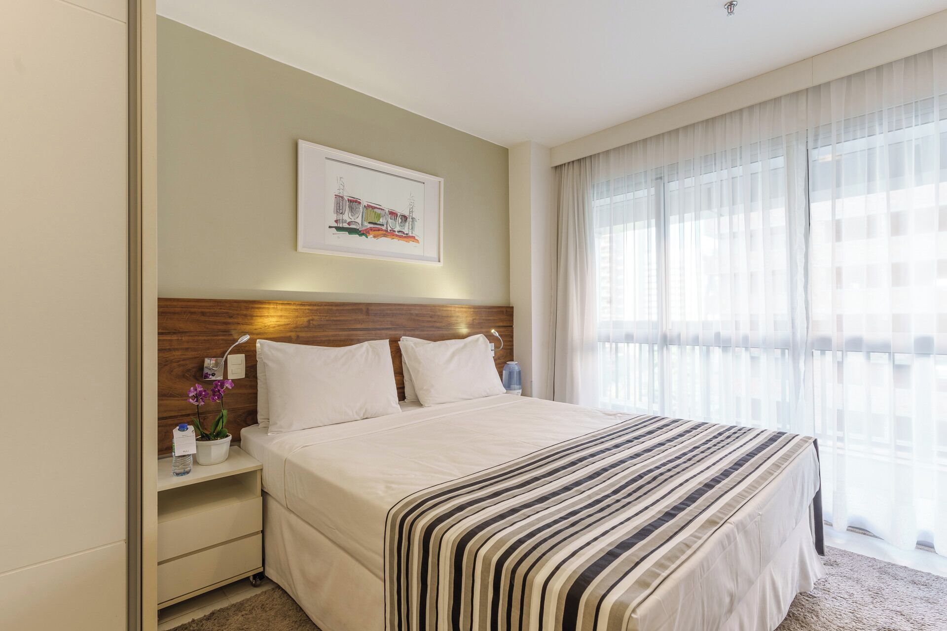 A hotel room with a large bed, a wooden headboard, a nightstand with a lamp, a framed picture on the wall, and a window with sheer curtains.