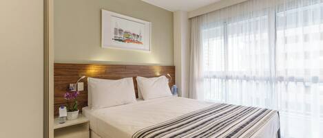 Ein Hotelzimmer mit einem großen Bett, einem Holzkopfbrett, einem Nachttisch mit Lampe, einem gerahmten Bild an der Wand und einem Fenster mit transparenten Vorhängen.