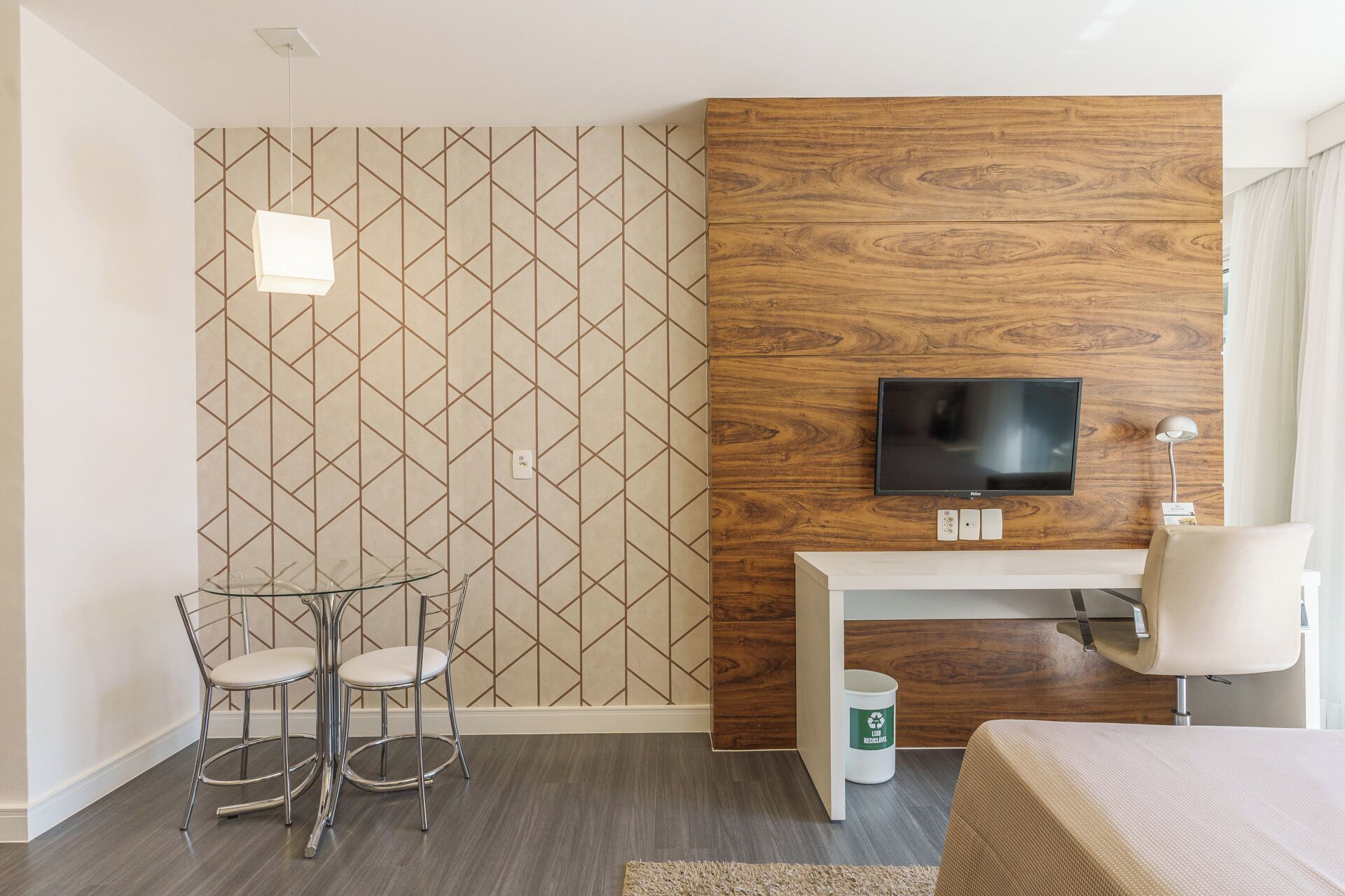 A modern hotel room with a geometric patterned wall, a wooden accent wall, a flat-screen TV mounted above a desk, two bar stools, a bed, and a recycling bin.