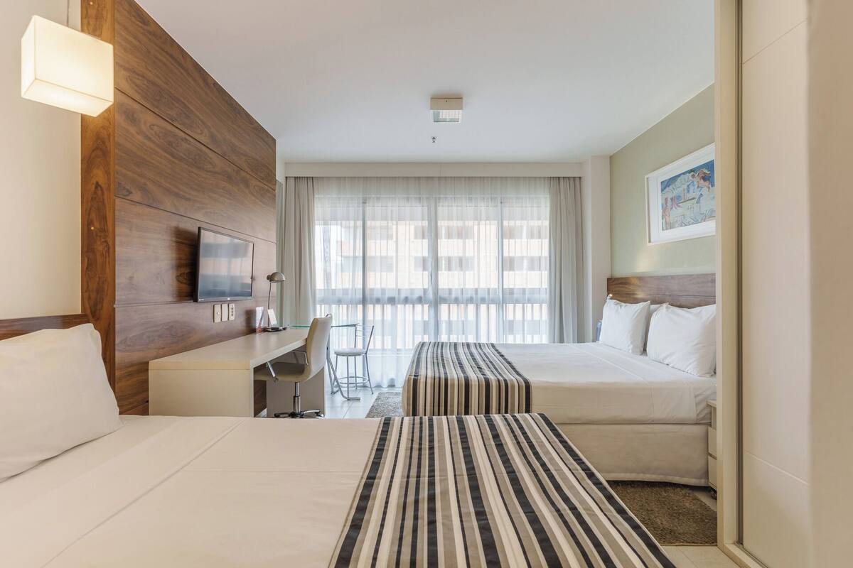 Une chambre d’hôtel comprenant un lit, un bureau avec une chaise, une télévision et une grande fenêtre avec des rideaux légers.