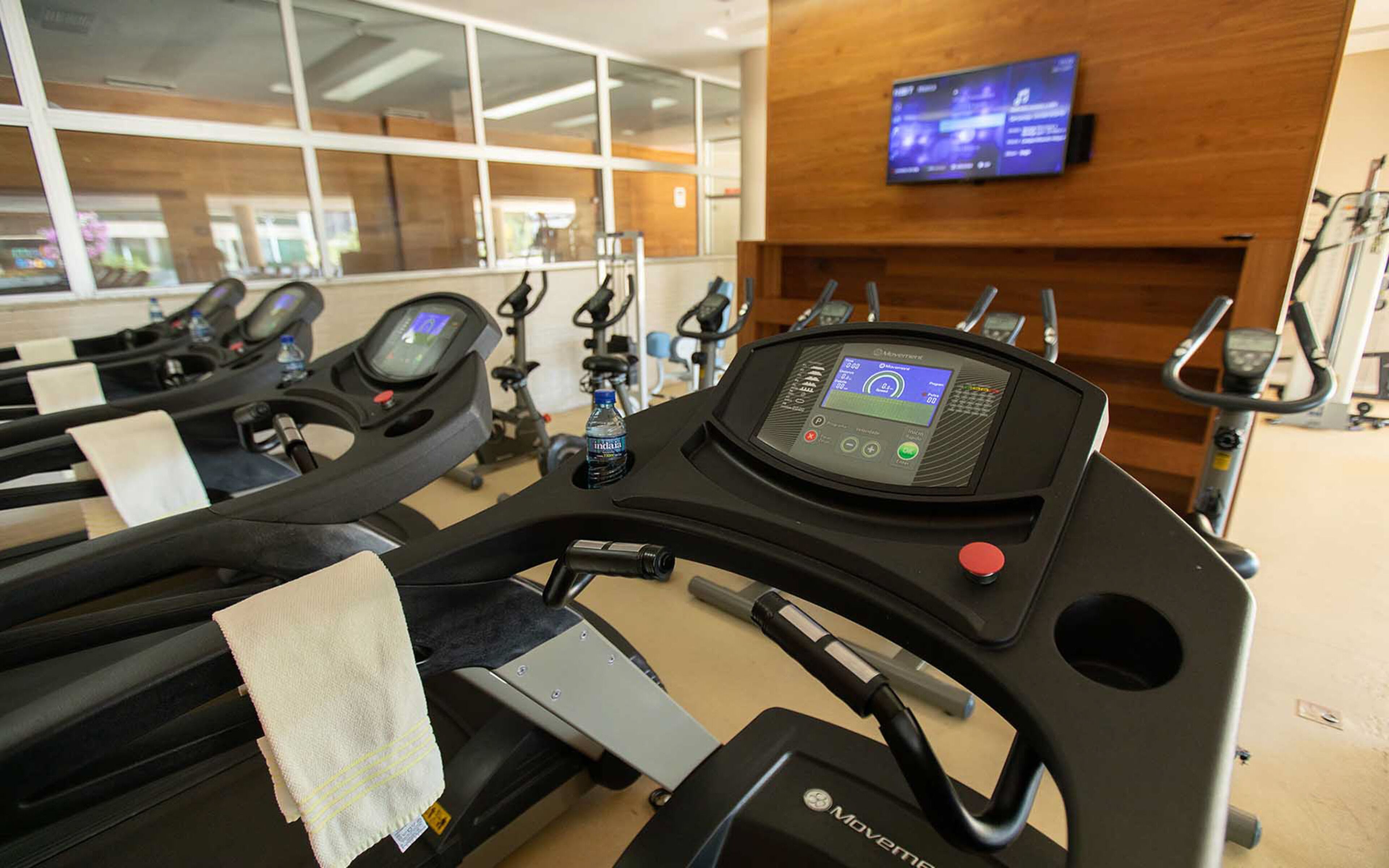A gym with treadmills, stationary bikes, and a TV screen displaying workout information.