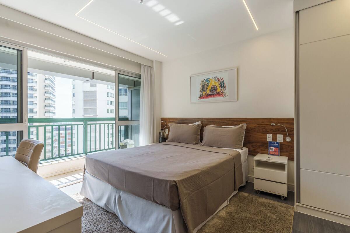 Une chambre d’hôtel avec un grand lit, un bureau, une chaise, un balcon donnant sur des immeubles et un tableau au mur.