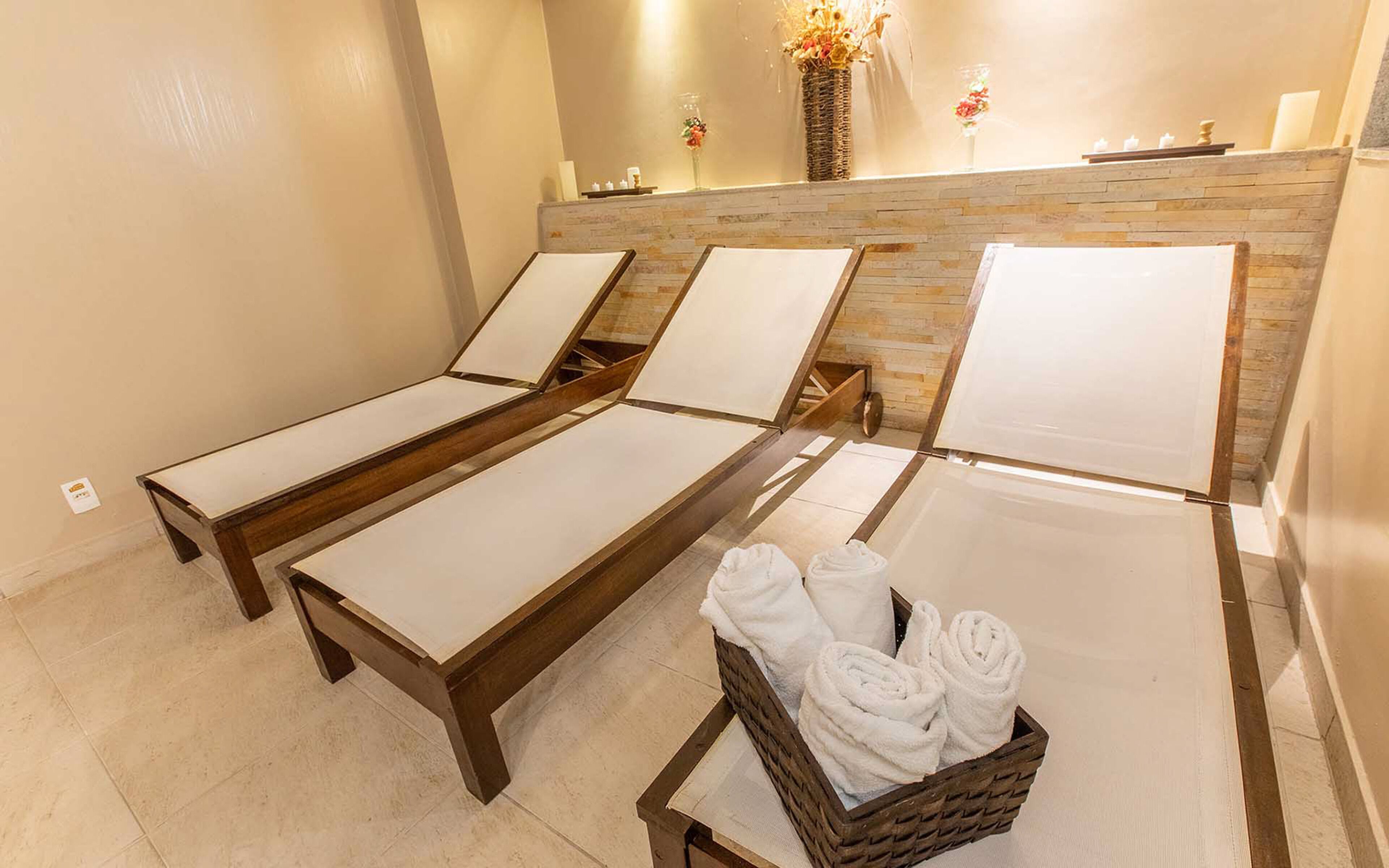 Un cuarto de spa con tres camastros de madera, una cesta de mimbre con toallas y una pared de piedra con un florero.