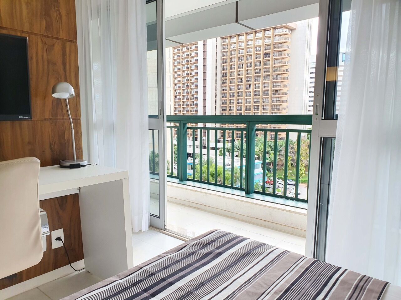 Une chambre moderne équipée d’un bureau, d’une chaise, d’un téléviseur fixé au mur et d’un balcon offrant une vue sur les immeubles et les espaces verts.