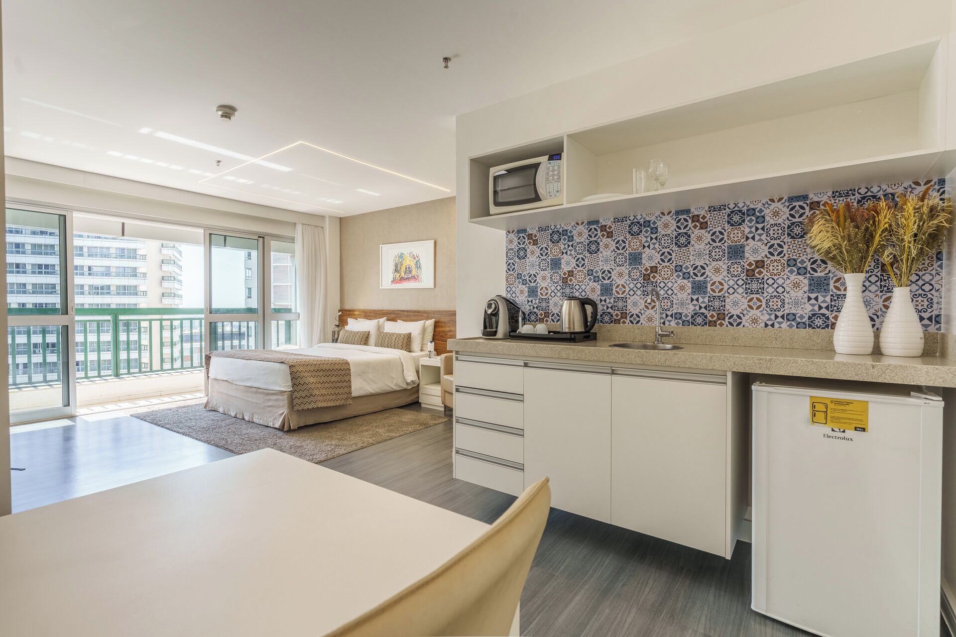 Habitación de hotel moderna con cama, kitchenette, zona de comedor y vistas a la ciudad.